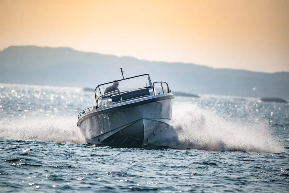 A speedboat splashes through water, driven by a person, with hills and a sunset in the background.